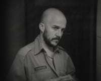 A bald, bearded man in a security uniform looks down thoughtfully in a dim room