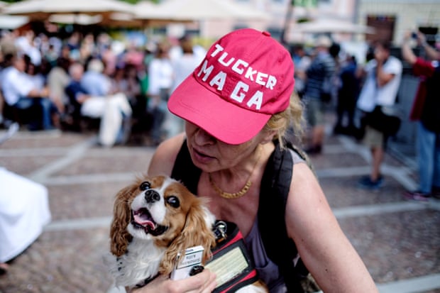 A woman wears a red hat with makeshift stickers spelling out 'Tucker Maga' as she looks down at a dog in her arms.