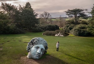 A person stands beside an enormous bronze head positioned on a lawn