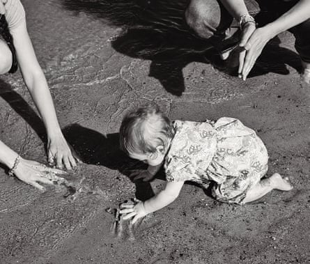 Black and white photo of a baby crawling in the sand on a beach, two women with her, hands in shot but faces out of view