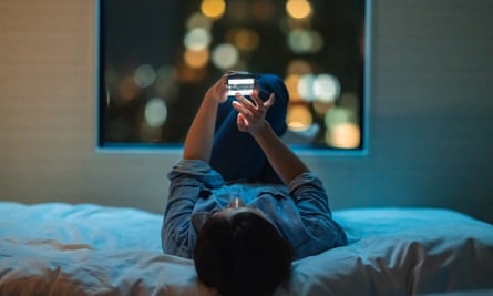 A woman is lying down on a bed and using a smartphone at night.