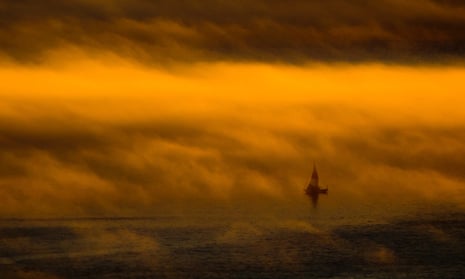 A sailboat as dense fog hangs over the water at sunset.