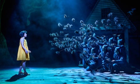 still image from stage performance of My Neighbour Totoro: a girl in a yellow dress and sandals stands to one side of the stage opposite a group of kneeling characters in hoods who are holding long sticks which have clumps of feathery vegetation at the end; the stage is lit in dramatic shades of blue