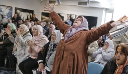 A woman, in coat and headscarf, raises her outstretched arms as other women sit applauding