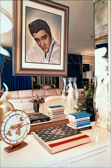 A framed portrait of Elvis Presley above a desk with a clock, a stars and stripes note book, and paperback books and a glass vase with a Greek statue inside it. A cream sofa and a silver tea set are mirrored in the background