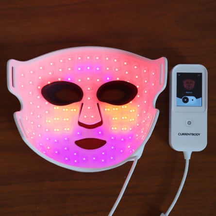 A glowing pink LED face mask lies beside a CurrentBody controller on a wooden surface