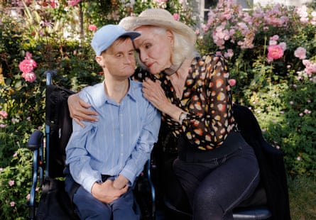 An older blond woman wearing a hat embraces a younger man with Down’s syndrome as he sits alongside her in a garden with rose bushes surrounding them.