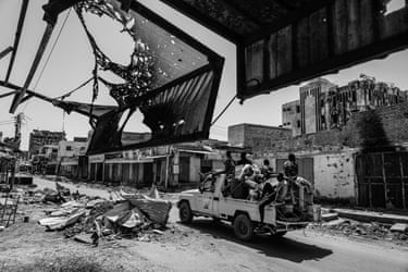 Black and white image of soldiers passing through a damaged market on a truck