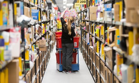 A fulfilment centre worker reaches up to shelves holding a scanner, beside a cart of red bins