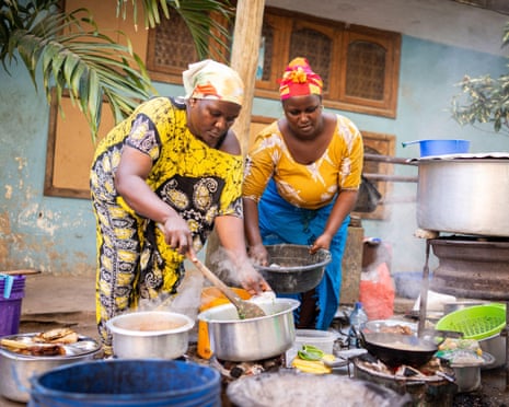 Two African women in colorful headscarves cook traditional food outdoors with multiple pots and pans
