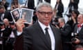 Director Mohammad Rasoulof, with short grey hair and wearing glasses and a shirt, tie and suit jacket, frowns as he holds up photos of his lead actors Missagh Zareh and Soheila Golestani, who were unable to leave Iran, with photographers behind him