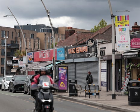 A motorcyclist with a learner plate rides past shops and parked cars in Ilford.
