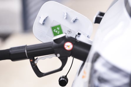 Close-up of a nozzle resembling a petrol pump nozzle in a car’s fuel filler. On the car’s fuel-cap flap there is a green diamond sign reading “H2 Gas”