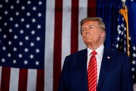 A man in a blue suit and red tie stands in front of American flags