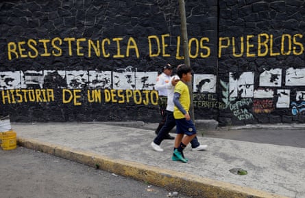 Two people walk past a wall painted with slogans in Spanish