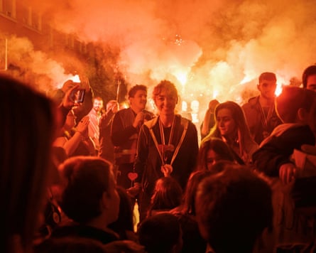 Sodium-orange flares light up a crowd of young protesters filling the frame as a young woman stands still and smiling at the camera in centre-frame