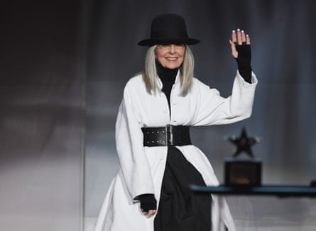 Diane Keaton accepting an award while wearing a white coat under a large black belt, black polo neck with overhand sleeves and large black hat