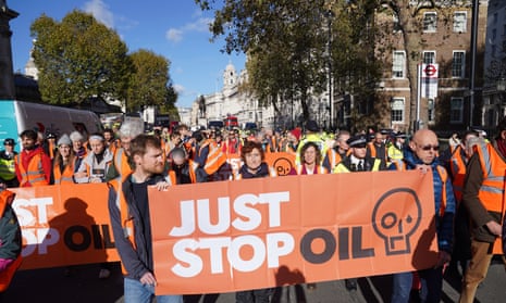 Just Stop Oil protesters take part in a walking protest in central London, 6 November 2023: they are wearing orange fluorescent tabards and holding bright orange banners as they block Whitehall. It is a fine, clear day with a blue sky, and trees and buildings can be seen in the background.