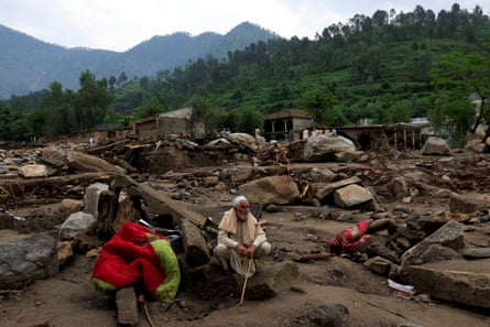 A man sits on and is surrounded by debris and rocks in a muddy landscape, with a green hill in the background