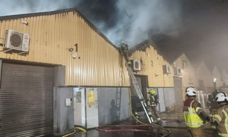 Warehouse, dark smoke and firefighters with hoses with one firefighter up a ladder