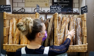 A baker holds a baguette inside a bakery in Paris, France, while wearing a facemask