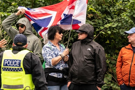 Protesters outside the Bell Hotel, Epping. A middle-aged woman holds a union jack flag; she stands between men wearing rain anoraks with their hoods up.