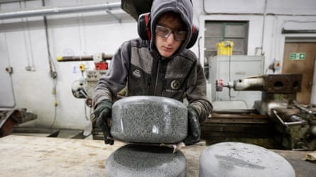 Man in a hoodie wearing ear protecters lifts a grey curling stone up in a factory