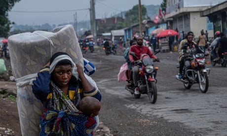 A woman with a child strapped to her front walks on the side of a road as two motorcycles pass by