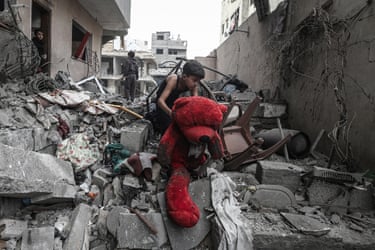 A boy lifts a large red teddy bear amid grey rubble and debris
