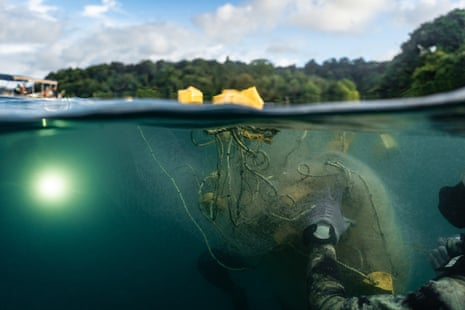 A diver rolls a net underwater. The sky and tree line can be seen above water, with a boat in the distance
