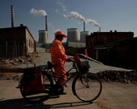 A man in orange hi-vis clothing walks while pushing his bicycle as chimneys belch smoke behind