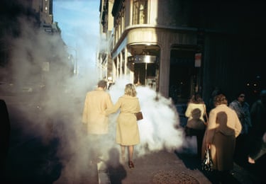 A group people all wearing beige-coloured coats walking past a manhole cover with steam spurting from it