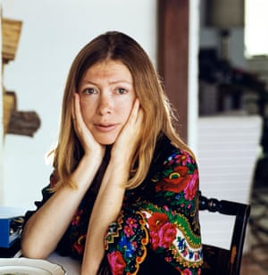 Joan Didion sits at a table wearing a shawl with a colorful floral print on a black background while cradling her face in her hands.