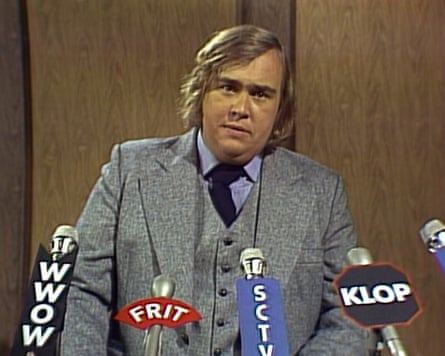 Candy (in character) gives a press conference, talking to microphones labelled with SCTV, KLOP, FRIT and WWOW