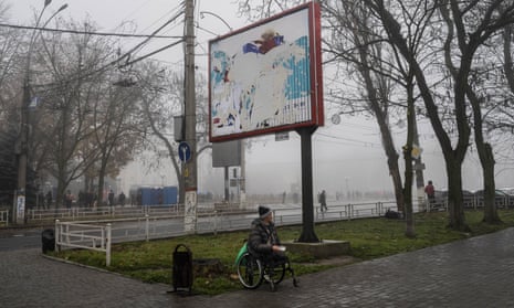 A man begs for alms under a billboard with visible remains of Russian posters.