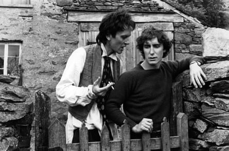 a black-and-white photo of two men in front of a stone cottage with a wooden picket fence.