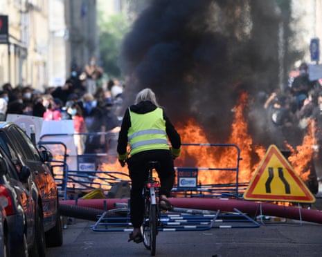 A cyclist in a yellow vest rides toward a burning barricade during a student protest