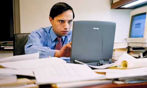Man sitting staring at a computer on a desk with lots of paper on it, looking stressed