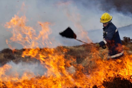 A firefighter wearing a heavy jacket and yellow helmet using a tool on a fire, with orange flames in the foreground