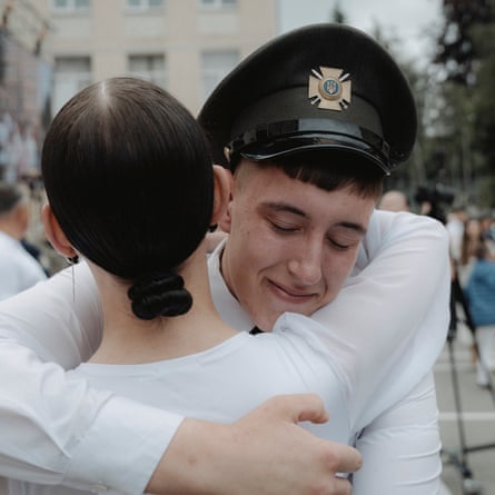A male cadet in a black peaked cap hugs a female cadet who has her hair tied back into a knot at the nape of her neck. They are both wearing white shirts.