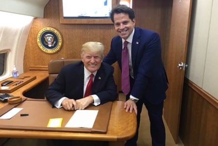 Donald Trump smiles as he sits at a desk with Scaramucci standing alongside him and smiling.