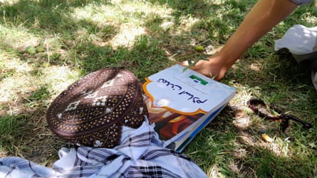 A hand holding a book with Arabic script, next to a traditional Hazara-type hat and prayer beads