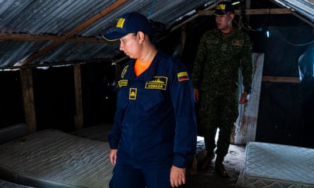 A woman in a dark blue uniform with yellow stitching followed by a man walk through a small wooden shack with a peaked roof. On both sides of them lay a single row of bare mattresses.