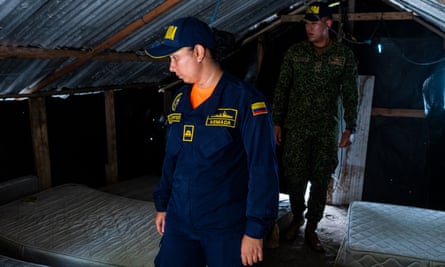 A woman in a dark blue uniform with yellow stitching followed by a man walk through a small wooden shack with a peaked roof. On both sides of them lay a single row of bare mattresses.