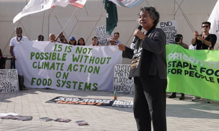 A brown-skinned, middle-aged Asian woman speaks into a microphone, while behind her two groups of people in a row hold banners, one of which says ‘1.5 is not possible without climate action on food systems.’
