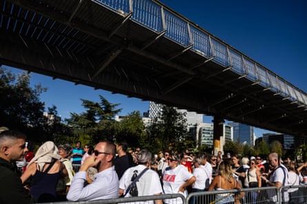 A line of people queueing in the sunshine underneath a bridge.