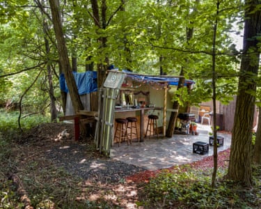 A shed made of various discarded building materials has a smart covered bar area attached, with a counter, three upholstered bar stools, and a barbecue alongside, plus an area covered in floor tiles to stand on