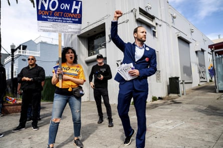 man with fist raised, wearing suit, next to woman holding sign saying ‘no on H’