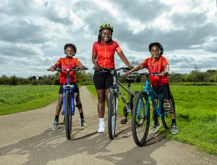 A woman and two young boys, all wearing red cycling tops, black cycling shorts and helmets, standing with bicycles on a track through countryside