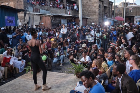 A woman wearing a black outfit with flat, tan-coloured lace-up shoes looks out at a crowd of people and cameras.